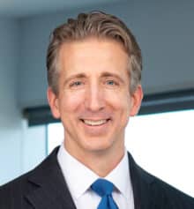 Professional headshot of Philadelphia community association and commercial construction attorney Matthew C. Collins standing in the Horn Williamson & Collins office. He is smiling and wearing a dark pinstriped suit, white dress shirt and bright blue tie, with short salt-and-pepper hair. Modern office windows and blue-gray finishes are visible in the background.