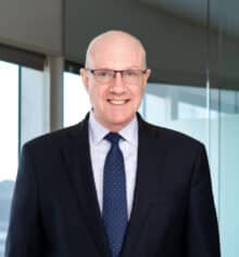 Professional headshot of Carter Williamson, a construction attorney at Horn Williamson, smiling and wearing a navy suit, light blue dress shirt and navy tie with small dots, with closely cropped gray hair and rectangular eyeglasses, standing in an office with large windows and a blurred Philadelphia cityscape in the background.
