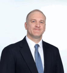Professional headshot of Patrick A. Costello, a Delaware and Philadelphia construction attorney and mediator. He is smiling, wearing a dark navy suit, white dress shirt, and light blue tie, posed against a light blue background.