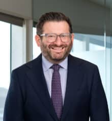 Professional headshot of Ryan Lockman, Managing Partner and residential construction defects attorney at Horn Williamson LLC, smiling and wearing a navy suit, light blue dress shirt and patterned tie, with short dark hair, a neatly trimmed beard and rectangular eyeglasses, standing in an office with large windows and a blurred Philadelphia cityscape in the background.