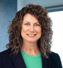 Professional headshot of Philadelphia commercial litigation and construction attorney Janesa Urbano standing in the Horn Williamson & Collins office. She is smiling and wearing a black blazer over a bright green blouse, with shoulder-length dark curly hair. Modern office windows and blue-gray finishes are visible in the background.