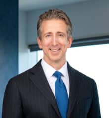 Professional headshot of Philadelphia community association and commercial construction attorney Matthew C. Collins standing in the Horn Williamson & Collins office. He is smiling and wearing a dark pinstriped suit, white dress shirt and bright blue tie, with short salt-and-pepper hair. Modern office windows and blue-gray finishes are visible in the background.