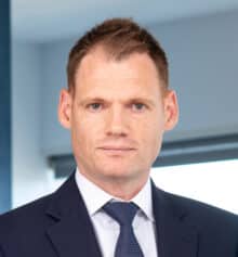 Professional headshot of Philadelphia commercial construction attorney Matthew H. Dempsey standing in the Horn Williamson & Collins office. He has short light brown hair with blond highlights and is wearing a navy suit, white dress shirt and blue patterned tie, looking at the camera with a neutral expression. Modern office windows and blue-gray finishes are visible in the background.