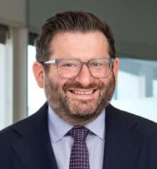 Professional headshot of Ryan Lockman, Managing Partner and residential construction defects attorney at Horn Williamson & Collins, smiling and wearing a navy suit, light blue dress shirt and patterned tie, with short dark hair, a neatly trimmed beard and rectangular eyeglasses, standing in an office with large windows and a blurred Philadelphia cityscape in the background.