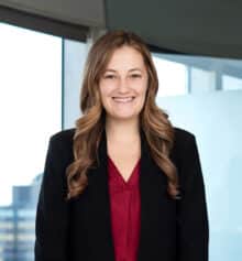 Professional headshot of Lauren McDowell, a residential construction defects attorney at Horn Williamson, smiling and wearing a black blazer over a red blouse, with long light brown wavy hair, standing in an office with large windows and a blurred Philadelphia cityscape in the background.