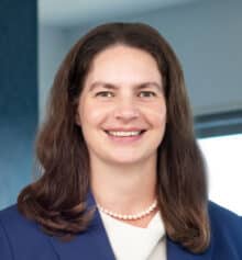 Professional headshot of Philadelphia commercial litigation and construction attorney Megan Keefe Shannon standing in the firm's Philadelphia office. She is smiling and wearing a blue blazer over a white blouse with a pearl necklace, with long brown hair. Modern office windows and blue-gray finishes are visible in the background.