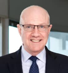 Professional headshot of Carter Williamson, a construction attorney at Horn Williamson, smiling and wearing a navy suit, light blue dress shirt and navy tie with small dots, with closely cropped gray hair and rectangular eyeglasses, standing in an office with large windows and a blurred Philadelphia cityscape in the background.